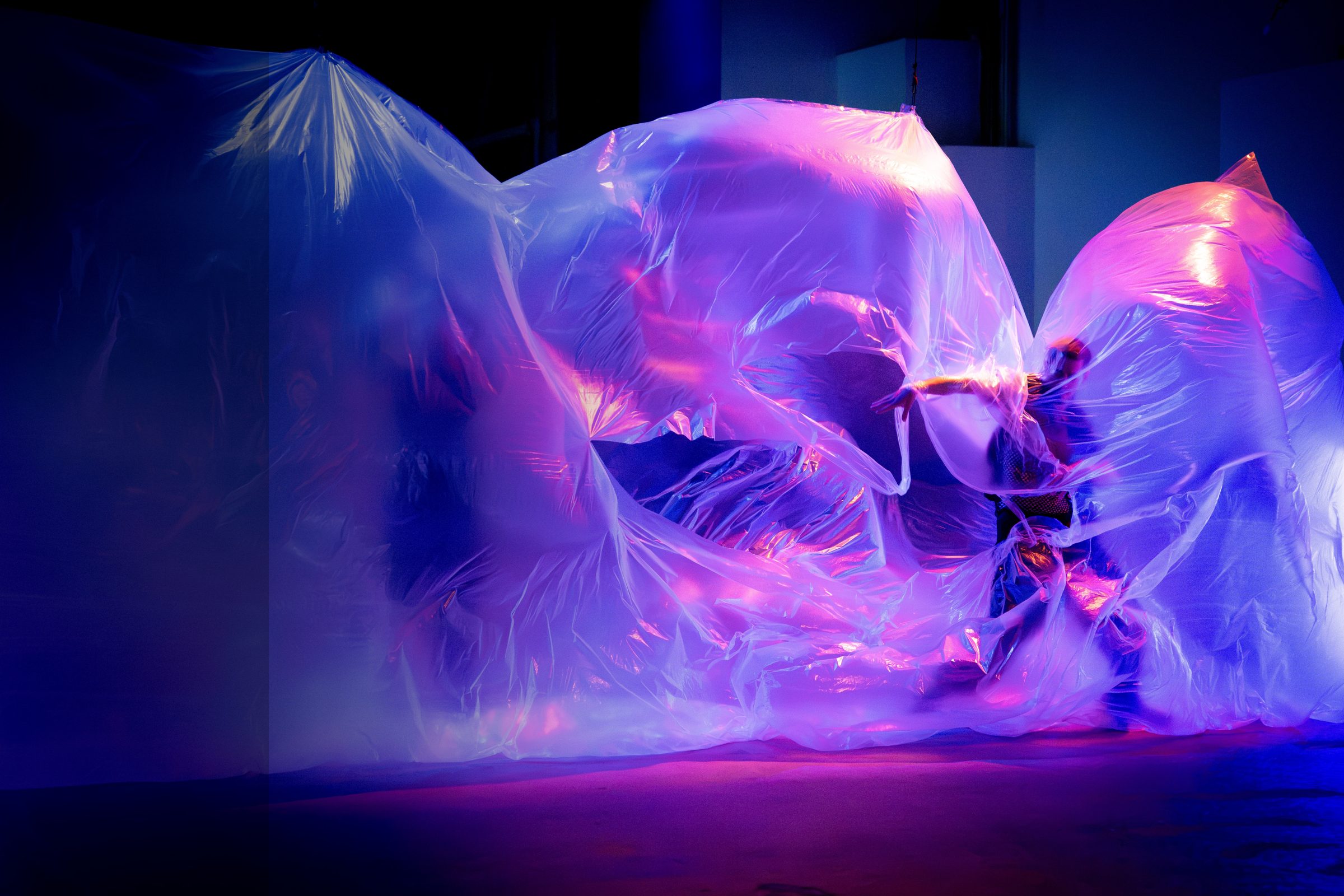 A dancer emerging from translucent sheeting lit in blue and magenta — The Still Point