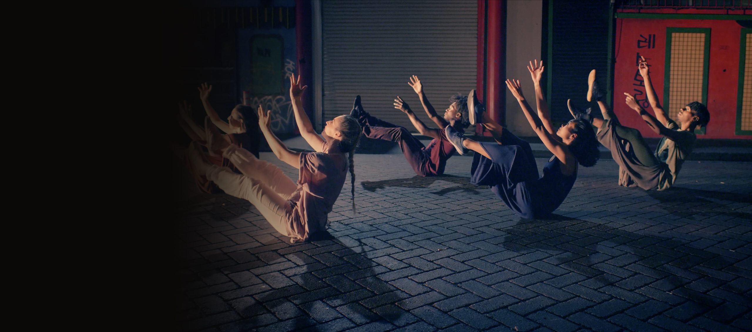 A row of dancers on a cobblestone street in v-hold, arms and legs raised toward sunset light, the leftmost figure emerging from shadow — Iris Company