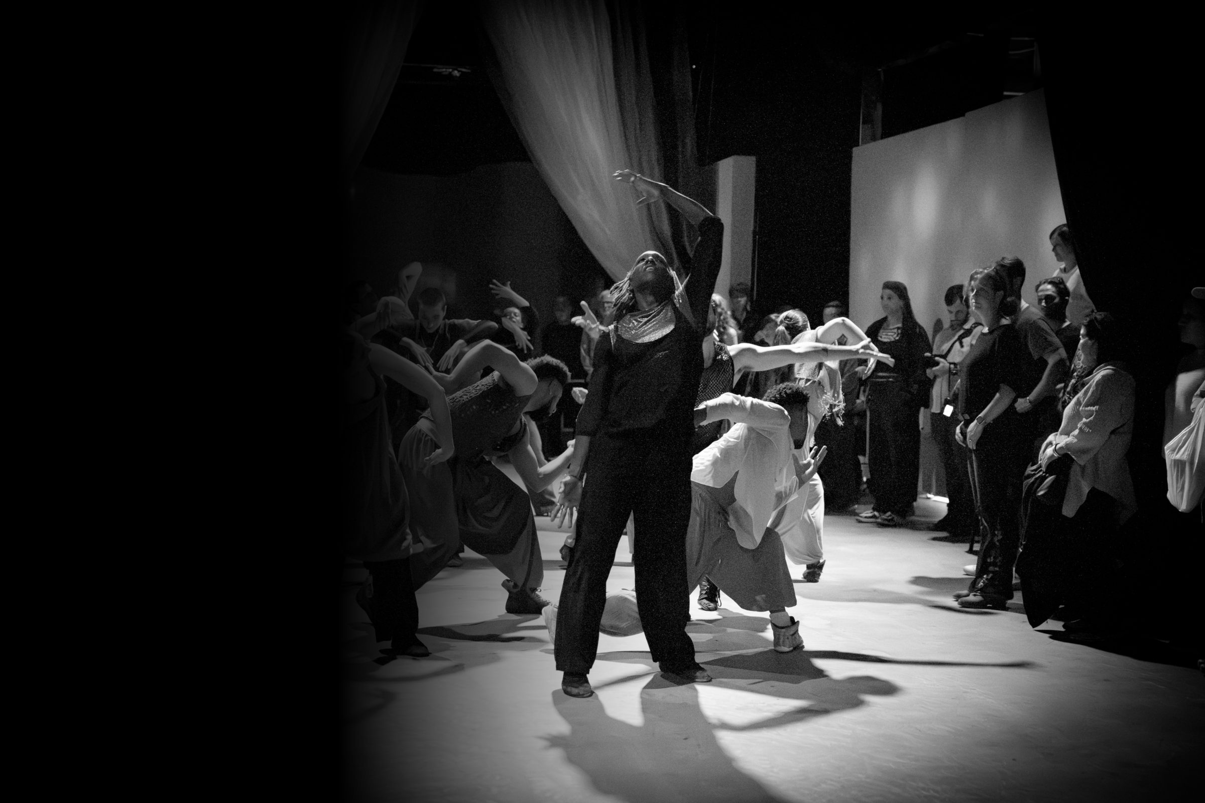 An ensemble of dancers in motion at the center of a gathered audience, in black and white — The Still Point