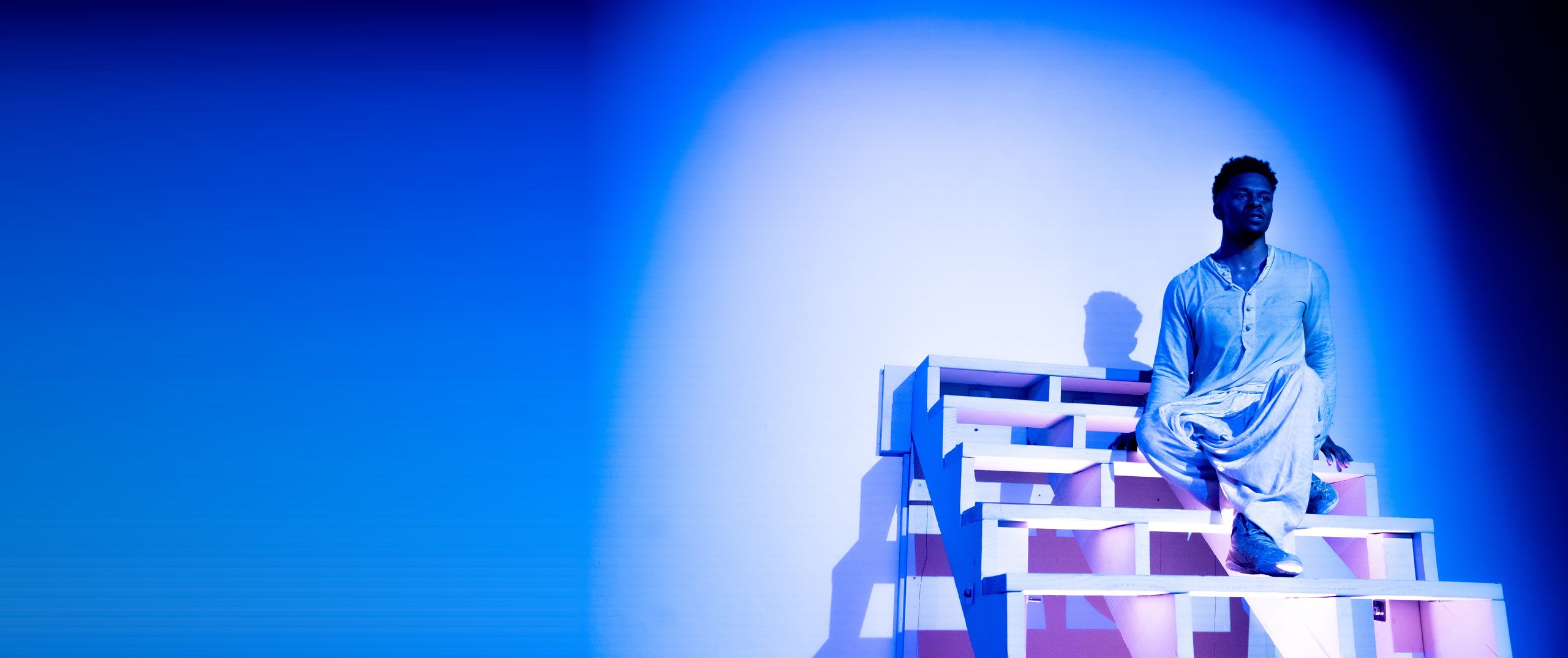 A dancer poised on a stepped set piece bathed in blue light, gazing outward — The Still Point