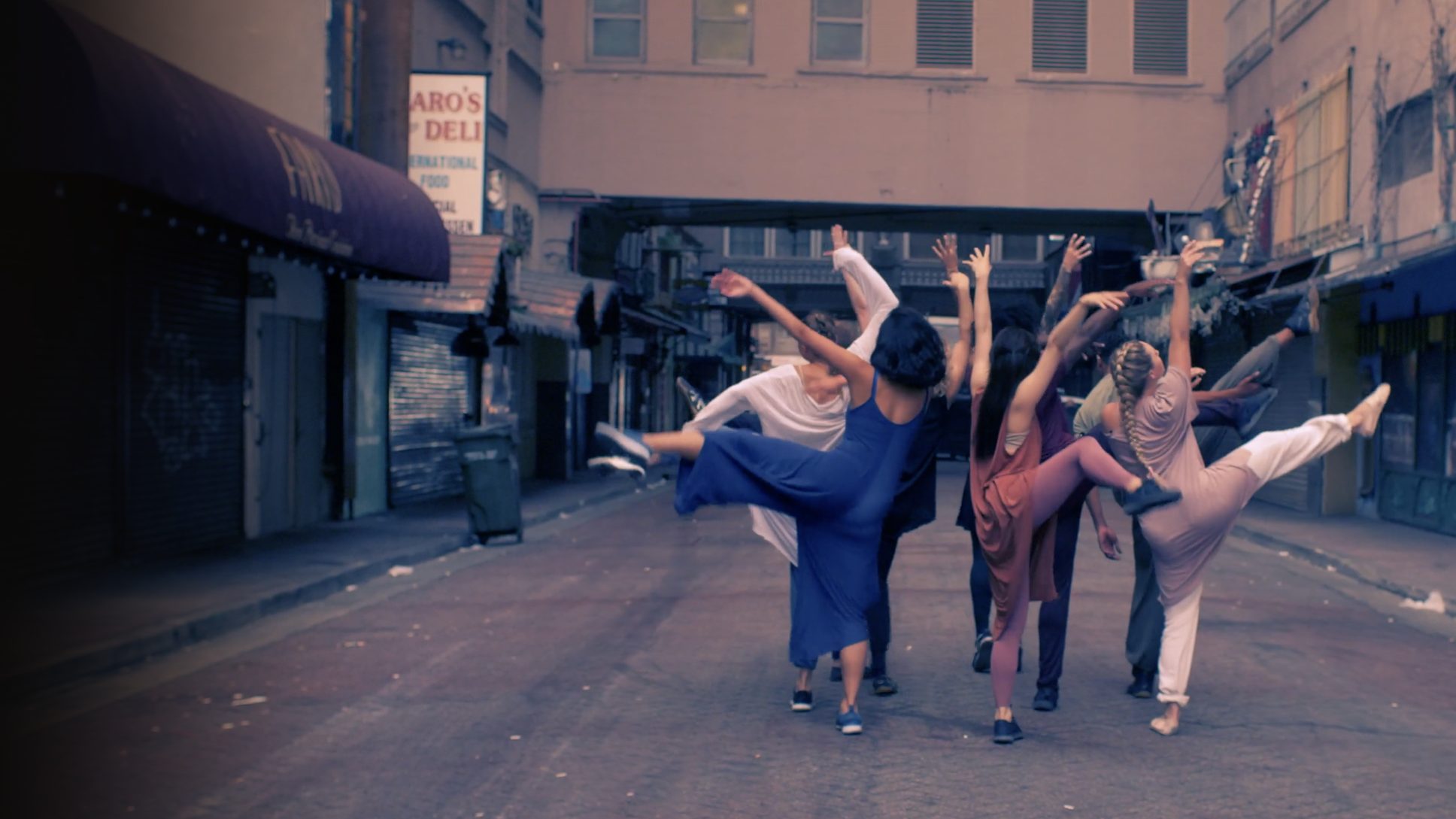 A cluster of dancers in a downtown alley extending arms and legs skyward in unison, the shopfronts fading into shadow — Iris Company